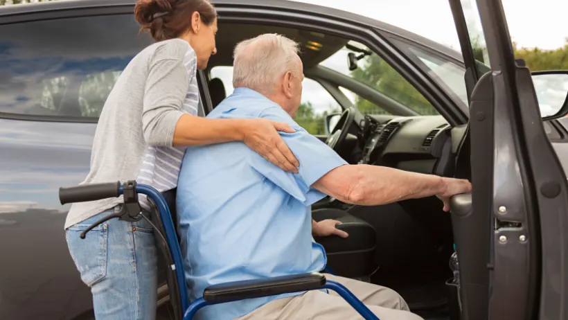 A woman helping an elderly man in a wheel chair get into the passenger seat of a car